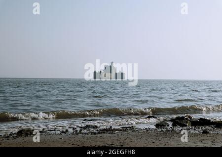 Leuchtturm im arabischen Meer, erbaut aus starken Felsen, die zwischen Wellen hoch stehen. Wellen an einem Sandstrand, der voller Kieselsteine und Felsen ist. Stockfoto