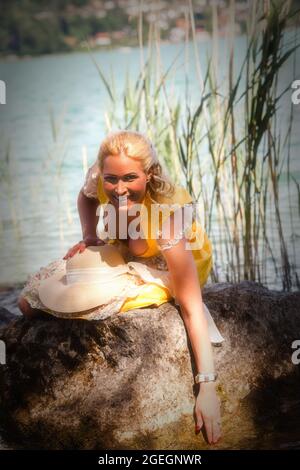 Elegante Frau mit Dirndl sitzt lächelnd auf einem Stein im See und hält eine Hand im Wasser. Stockfoto