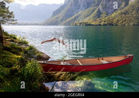 Eine Frau, die in der Nähe des wunderschönen roten Kanus auf einem abgeschiedenen Teil des Bohinjsees in Slowenien ins Wasser springt Stockfoto