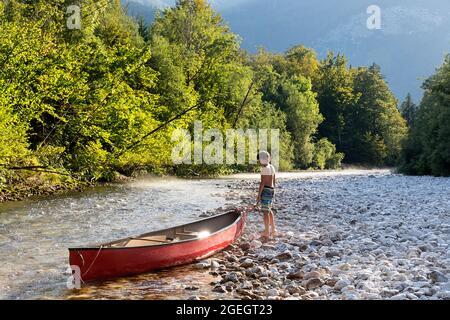 Ein Junge, der sich um ein wunderschönes rotes Kanu auf dem Sava Bohinjka River kümmert, kurz vor dem Bohinjsee, Slowenien Stockfoto