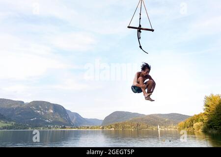 Ein Junge, der an einem wunderschönen Sommertag auf dem Bohinjsee in Slowenien von einer Baumschaukel springt Stockfoto