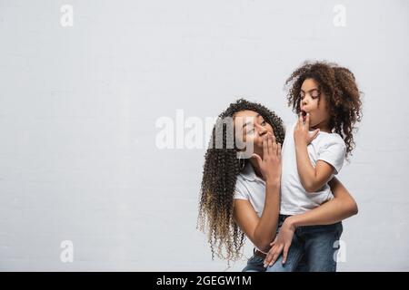 Verblüffte afroamerikanische Mutter mit Kind, das den Mund mit der Hand bedeckt und sich auf grau anschaut Stockfoto