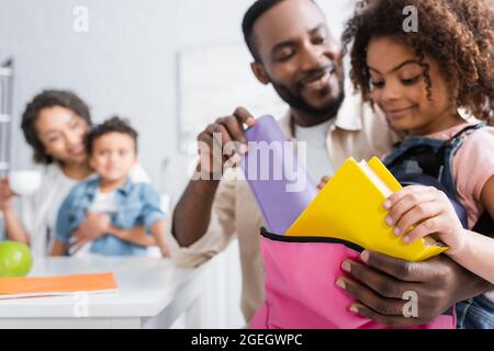 afroamerikanischer Mann hilft Tochter Rucksack in der Nähe verschwommen Familie in der Küche packen Stockfoto