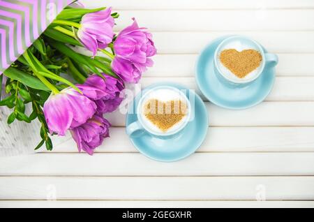 Zwei Tassen Cappuccino mit einem Herzförmigen Symbol und lila Tulpen auf einer hölzernen Hintergrund. Stockfoto