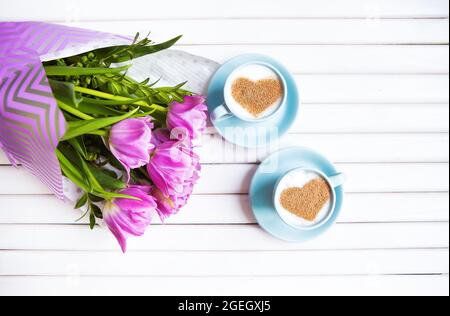Zwei Tassen Cappuccino mit einem Herzförmigen Symbol und lila Tulpen auf einer hölzernen Hintergrund. Stockfoto