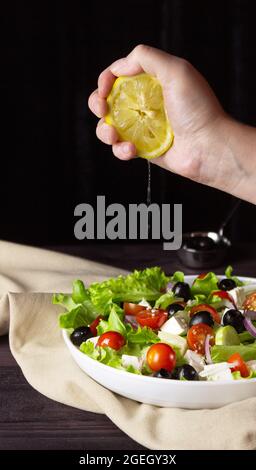 Griechischer Salat auf einem Teller auf dunklem Grund. Gesundes vegetarisches Essen. Die Hand drückt den Zitronensaft aus. Feta-Käse, Salatblätter, kirschtomat Stockfoto