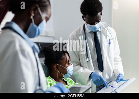 Medizinisches Team von afroamerikanischen Ärzten, das jungen Patienten, die im Krankenhausbett sitzen, Gesundheitsberatung gibt. Afro-Leute überprüfen Diagnose für kranke Mädchen mit Genesung Ausrüstung Stockfoto