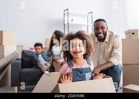 Aufgeregt afroamerikanischen Mädchen lachen in Karton in der Nähe verschwommen Papa und Mutter mit Bruder Stockfoto