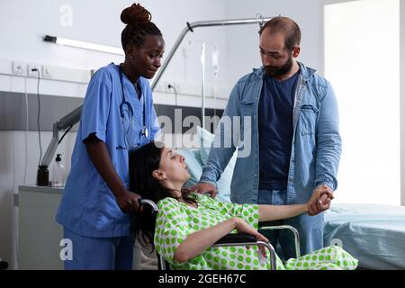 Eine Schwangere im Rollstuhl erhält Hilfe von Krankenschwester und Ehemann, während sie sich auf die Geburt im Entbindungszimmer der Klinik vorbereitet. Medizinische Assistentin hilft dem Paar, das ein Baby erwartet Stockfoto