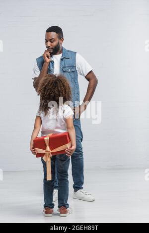 Rückansicht eines afroamerikanischen Kindes, das hinter dem Rücken eine rote Geschenkbox in der Nähe eines nachdenklichen Vaters auf Grau hält Stockfoto