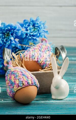 Osterkomposition mit Eiern in Strickmützen, blauen Blumen und dekorativem Kaninchen auf einem Holztisch. Stockfoto