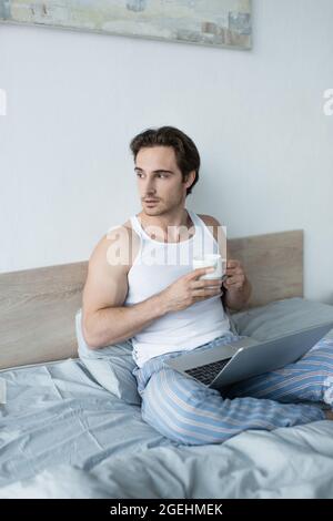 Junger Mann schaut weg, während er mit einem Laptop und einer Tasse Kaffee im Bett sitzt Stockfoto