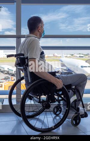 Junger Mann mit Schutzmaske im Rollstuhl am Flughafen und Blick aus dem Fenster Stockfoto