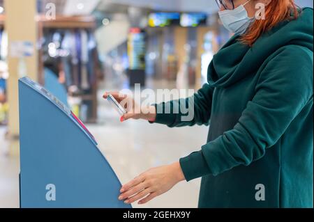 Eine Frau in einer Maske scannt den qr-Code von ihrem Smartphone aus. Ein Mädchen nutzt am Flughafen einen Self-Service-Automaten, um für einen Flug einzuchecken und Tickets auszudrucken Stockfoto