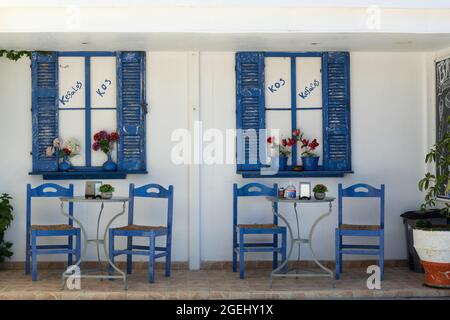 Ziemlich traditionelle blaue und weiße Fensterläden und Stühle vor einer Taverne in Kamari, Kefalos, Kos, Griechenland Stockfoto