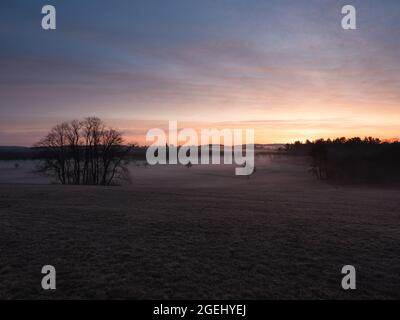 Sonnenaufgang über einem Bauernhof in Massachusetts. Dieses Heufeld ist voller Nebel und einsamer Bäume. Der Morgenhimmel hat ein paar Wolken und viele Farben. Stockfoto