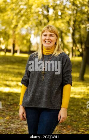 Unschärfe lächelnde blonde 40er Frau im gelben Herbst Park stehen. Frohe, schöne Dame. Frauen tragen grauen Pullover, gelben Rollkragen, Anhänger, Jeans. Stockfoto