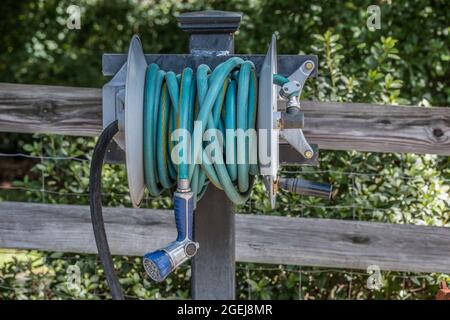 Robuster Gartenschlauch mit einem Handsprüher, der auf einer Metallrolle befestigt ist, die auf einem Pfosten gegen den Zaun in einem Garten für Wasser sup montiert ist Stockfoto