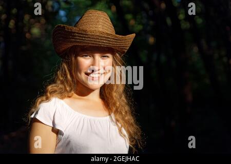 Nahaufnahme Porträt eines jungen Langhaar-Mädchens in einem Strohhut in hellem Sonnenlicht vor verschwommenem dunklen Hintergrund. Copyspace. Stockfoto