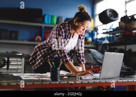 Mixed-Race-Automechanikerin, die am Schreibtisch steht und in der Werkstatt einen Laptop benutzt Stockfoto