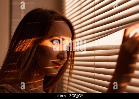 Nahaufnahme eines Mädchens, das durch die Jalousien im warmen Licht der untergehenden Sonne aus dem Fenster schaut. Geringer Fokus. Stockfoto