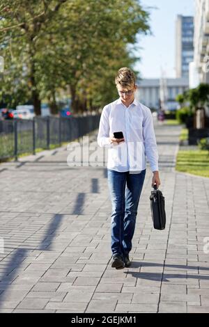 Junger Mann, der die Straße entlang läuft, mit einem Telefon in der Hand und einer Aktentasche in der anderen Hand. Geringer Fokus. Stockfoto