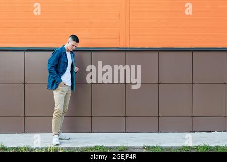Der junge Mann in einer blauen Jeanjacke und einer beigen Hose steht nachdenklich gegen die Wand. Copyspace. Stockfoto