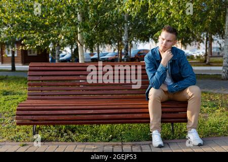 Der junge Mann sitzt mit nachdenklicher Optik auf einer Parkbank. Copyspace. Stockfoto