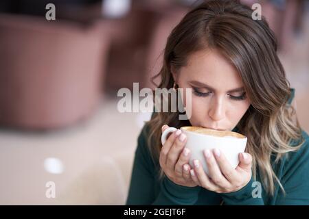 Ein glückliches schönes Mädchen mit einer schönen Frisur, hält eine Tasse Kaffee, in einem grünen Rollkragen, sitzt in einem Café, Kaffee trinken. Hochwertige Fotos Stockfoto