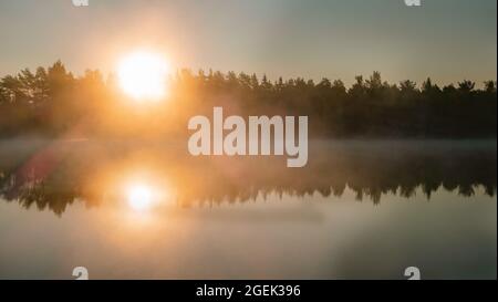 Landschaftlich neblig glühender Sonnenaufgang über dem See mit großer orangefarbener Sonne, Sonnenstrahlen steigen von der Wasseroberfläche Nebelküste mit Bäumen auf, Spiegelreflexion im See su Stockfoto