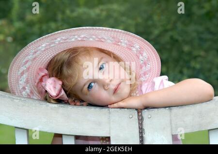 Entzückend in einem rosa Hut lehnt das kleine Mädchen ihren Kopf gegen ein rustikales Holztor. Sie hat einen engelhaften Ausdruck von pur Unschuld. Stockfoto