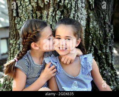 Zwei Schwestern sitzen draußen neben einem Baum. Sie zeigen ihre Freundschaft und Zuneigung füreinander, indem sie sich umarmen und einen weichen Kuss auf die Wange nehmen. Stockfoto