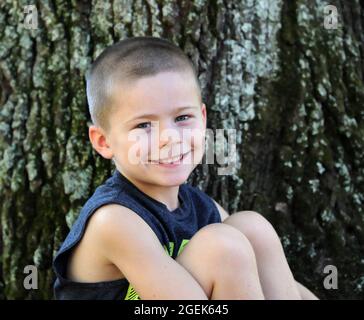 Das jungenhafte Grinsen breitet sich über dieses Gesicht der kleinen Jungen aus. Er sitzt draußen neben einem Baum und schaut auf die Kamera. Stockfoto