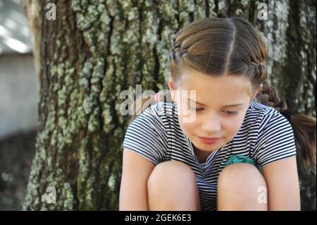 Kleines Mädchen ist in Gedanken verloren. Sie sitzt ganz allein neben einer Eiche. Sie schaut nach unten. Stockfoto