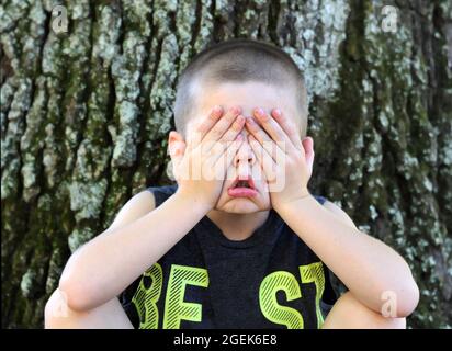 Der kleine Junge sitzt draußen gegen einen Baum und blitzen die Augen, damit er nicht hinsehen kann. Seine Hände quetschen seine Augen zu und schrubben ihm in einer komischen Ausrudung ins Gesicht Stockfoto