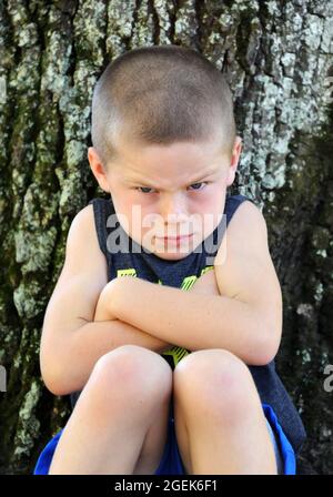 Kleiner Junge lehnt sich an einen Baum draußen und pout. Er hat die Arme gefaltet und die Augenbrauen gekreuzt. Stockfoto