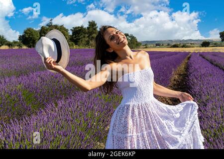 Porträt einer glücklichen jungen Frau, die mit ihrem Hut in einem blühenden Lavendelfeld spielt. Ihr weißes Kleid hebt sich von der violetten Farbe des Lavendelflos ab Stockfoto