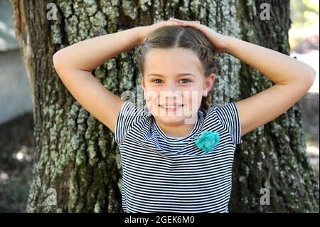 Kleines Mädchen sitzt neben einer Eiche. Sie hat ihre Hände hinter ihrem Kopf angehoben und lehnt sich an den Baumstamm. Stockfoto