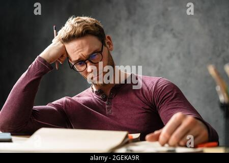 Das Porträt eines müden Mannes, der seinen Kopf mit der Hand stillt, während er mit Papieren im Studio am Tisch sitzt Stockfoto