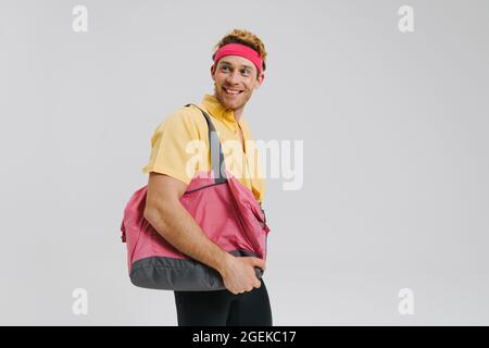 Lächelnder junger, weißer, schwuler Mann mit Stirnband, der über grauem Wandhintergrund steht und eine Sporttasche trägt Stockfoto