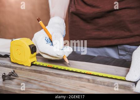 Exakt gemessen. Nahaufnahme eines jungen männlichen Zimmermanns in Handschuhen, der mit gelbem Maßband Messungen auf der Holzdiele anfertigte. Stockfoto