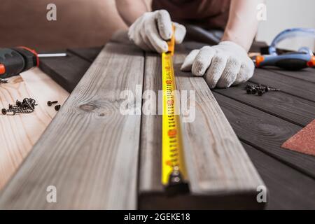 Exakt gemessen. Nahaufnahme eines jungen männlichen Zimmermanns in Handschuhen, der mit gelbem Maßband Messungen auf der Holzdiele anfertigte. Stockfoto