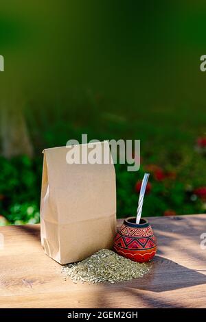 Eine rustikale Packung braunes Papier mit einem traditionellen Mate aus Kalabash über einem Holztisch mit Yerba Mate darüber verstreut. Vertikales Format. Stockfoto