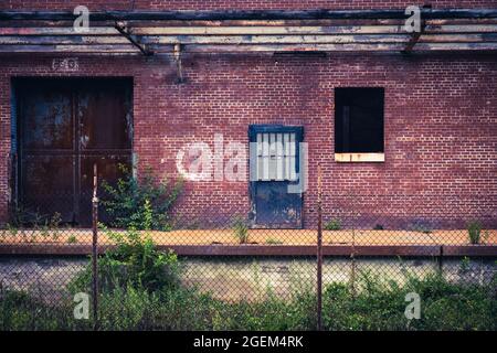 Alte Fabriktüren, die in einer Ziegelwand auf einer verlassenen Ladedock mit einem Kettengliederzaun im Vordergrund stehen Stockfoto