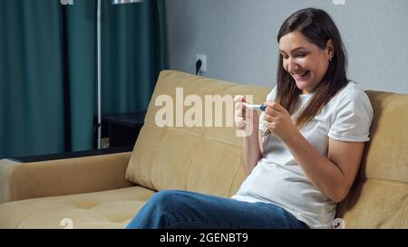 Glückliche Frau, die einen Schwangerschaftstest in den Händen hält, die auf dem Sofa sitzen Stockfoto