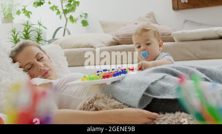 Müde Mutter schlief ein, während ihr Kind Baby Sohn mit bunten Lernspielzeug spielt, zu Hause zwischen Kissen liegt, gesund und gepflegt wachsen Stockfoto