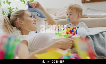 Glücklich lächelnde Mutter spielt mit ihrem schönen Baby, mit bunten Lernspielzeug, sitzt zu Hause zwischen Kissen und Tiere Plüsch, gesund und Stockfoto