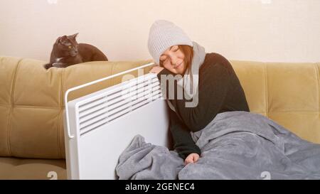 Die gefrorene Dame lehnt sich an die Heizung, die neben der Katze auf dem weichen Sofa sitzt Stockfoto