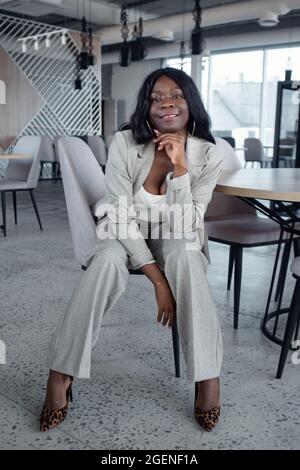 Die junge afroamerikanische Frau im Business-Anzug sitzt auf einem Stuhl Stockfoto