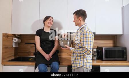 Eine junge Frau sitzt auf dem Tisch, ein Mann steht in der Nähe und mischte Teig mit einem Schneebesen Stockfoto
